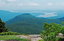 A view from a rocky mountain ledge over a lower mountain to a distant, winding lake