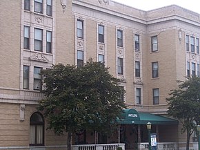 Front façade of the four-floor Antlers Hotel. constructed of tan brick with ornamental stone and terra cotta features in the Renaissance Revival style, and a porch surrounded by opposing bays. Both the porch and windows have balusters and classic detailing.