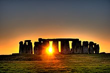 Sun shining through row of upright standing stones with other stones horizontally on the top.