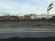 Two locomotives parked next to a building, with more tracks in the foreground along with a signal gantry.