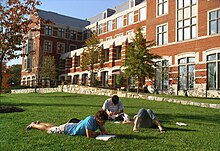 Three young adults lie on grass reading books in front of a brick building with many windows