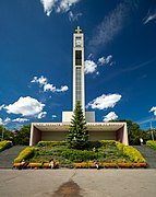 Une église moderne avec en son centre un impressionnant clocher parallélépipédique surmonté d'une croix d'or ; le corps de l'édifice monte en escalier le long de la pente du terrain ; de la pelouse et des fleurs au premier plan.