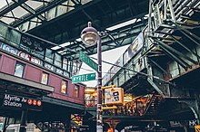 Von der Straße aus nach oben blickend. Vorne in der Mitte ein Ampel-Laternenmast mit den Straßenschildern MYRTLE AV und BROADWAY. Links in blutrot lackiertem Blech ein eingeschossiger, aufgeständerter Stationsbau, der mit schmalen hohen Fenstern an einen alten Eisenbahnwagen erinnert. Darunter ein moderner Wegweiser mit den runden, braunen bzw. orangenen Emblemen der Linien J, Z und M sowie dem Stationsnamen „Myrtle Av Station“. In der Mitte im Hintergrund eine vergitterte, beleuchtete Verteilerbrücke mit Menschen, rechts diagonal in Bild hineinlaufend ein Hochbahntragwerk mit silbrigem Zug darauf. Ganz oben im Vordergrund wiederum querend ein dunkelgrünes Hochbahntragwerk, das aber stillgelegt ist. Die dominierenden Farben sind industrielle Grün-, Rost- und warme Lichtorangetöne mit einzelnen anderen Farbakzenten, Stickern, Graffititags.