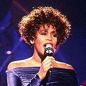A black woman short curly brown hair wearing a purple dress sings to a microphone
