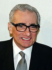 A Caucasian man in his 70s smiling. He is wearing black framed glasses and a tie with a black suit over a white shirt. Behind him is a white background