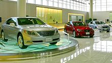 Một showroom trưng bày ba chiếc sedan, chiếc gần nhất nằm trên sàn xoay bằng kính, sau đó là trước quầy lễ tân và cuối cùng là cửa sổ.