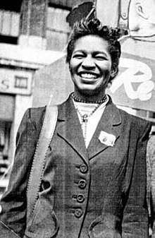 A woman with medium-dark skin tone and short black hair smiles at the camera. She wears a neat buttoned-up jacket and carries a shoulder bag with a large strap.