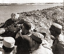 Un homme en costume regarde au-delà d'un cours d'eau avec des jumelles abrité derrière une butte de terre. Trois hommes en uniforme se tiennent derrière lui.