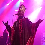 Tidig Papa Emeritus I Glitterkantig vit mitra med ghostkrucifix i svart, samt svart mässhake med glitterkantiga röda remsor längs kanten och centerlinjen pyntade med svarta nedåtvända krucifix.