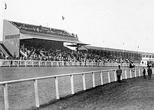 Black and white photo of horses running around a dirt track with a grandstand full of spectators in the background