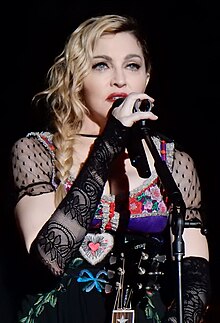A closeup photo of Madonna with shoulder-length wavy blonde hair, wearing a colorful, low-cut blouse, holding a microphone to her mouth with her right hand.