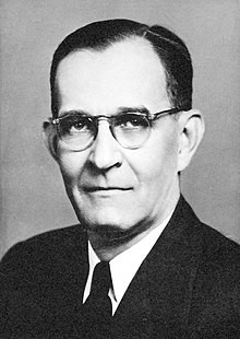 Black and white photograph of a white man with dark hair, wearing a suit and glasses