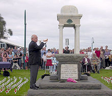 Ata o se faamanatu ANZAC ma se tagata taaalo i se bugle matutua. o loo nonofo laina o tagata i tua o le faamanatuga. E toatele laiti satauro paepae ma poppies mumu ua pipii i le vao i le laina i le itu o le fanua faamanatu.