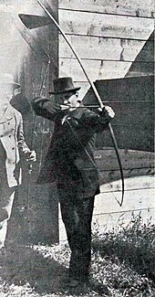 Photo d'un homme en costume et haut de forme s'apprêtant à décocher une flèche avec un arc.