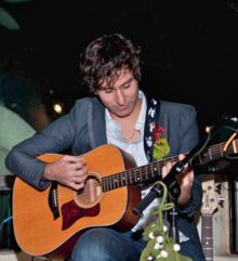 A dark-haired man sitting down playing guitar.