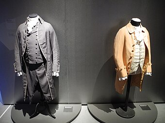 Two suits hung on valet stands in a display exhibit, a black one on the left and a tan one on the right