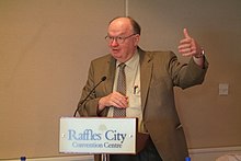 MacPherson, a mature Caucasian man wearing a suit and tie, speaking at a conference. Gesturing with one hand, he stands behind a podium bearing a convention centre logo.