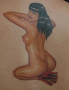 Detail tetování s Bettie Page na lidském těle; nahá Bettie Page klečí směrem od diváka, ke kterému se ale otáčí hlavou a smyslně se na něj dívá; má zvednuté pokrčené paže, černé rozpuštěné vlasy, opálenou pleť a červené lodičky na nohou