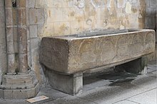 Photographie d'un sarcophage en granite avec un décor d'arcades en plein cintre en bas-relief sur le côté, et une croix ancrée à sa tête.