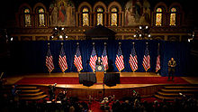 A speaker at a podium in the center of a stage decorated with nine American flags and a red carpet.