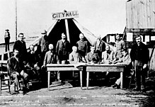 Men standing and sitting around two tables, facing the câmera. A large tent behind them has a wooden sign that reads "City Hall"