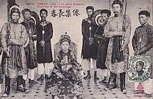 Photo ancienne représentant un jeune enfant asiatique, en habit de cour richement décoré, assis sur un trône. Sept homme asiatiques, en habit traditionnel, se tiennent debout à ses côtés.
