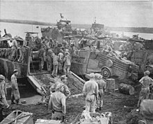Military personnel unloading stores and vehicles from landing craft on a beach