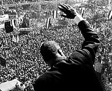 Photographie du dos d'un homme saluant une foule depuis le sommet d'un bâtiment