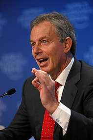 Tony Blair i mörk kavaj, vit skjorta och röd slips. Han talar och gestikulerar energiskt.