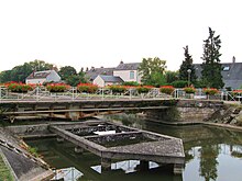 Photo couleurs. Le pont tournant de Combleux est vu de côté. Il est constitué d’une poutre métallique ajourée reposant sur une articulation. Au droit de celle-ci on devine l’emplacement du mécanisme faisant pivoter l’ouvrage. Devant se situe une sorte d’estacade. Les garde-corps du pont portent des pots de fleurs rouges.