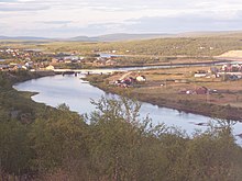 Foto eines Flusses, an beiden Seiten des Flusses erstreckt sich ein Dorf. Über den Fluss führt eine Straßenbrücke.