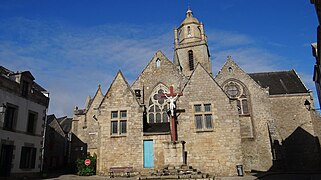 Photo d’une église en pierre, en arrière-plan d'une rue bordée de maisons couvertes d’ardoises.