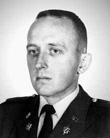 Head of a solemn-faced young man with short hair wearing a dark military jacket with pins on the lapels over a shirt and tie.