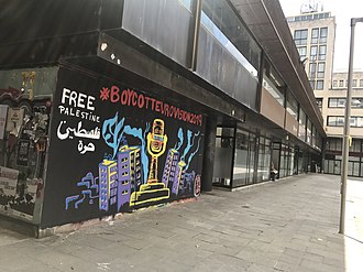 A painted mural on a wall on a street in Girona, Spain: the Eurovision trophy appears covered in barbed wire surrounded by tower blocks, with the words "#BoycottEurovision2019" above, and "Free Palestine" in English and Arabic to the top left