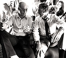 Photograph of two seated men having a conversation in a crowded busy room; the man on the left is giving "the finger" to the camera