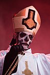 Sen Papa Emeritus I Guldig mitra med brett vitt kors försett med svartkantig ghostkrucifix i guld, samt vit mässhake med svart insida utsmyckad med "Ghostkrucifix" i guld längs ytterkanten och över ytterbröstet.