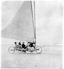 Photographie noir et blanc de quatre personnes à bord d'un char à voile sur une plage.