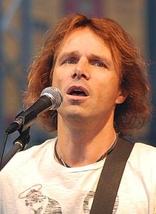 Janek Ledecký seen close-up in front of a microphone, holding a guitar (not in picture), looking to one side of the camera