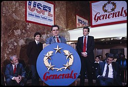 Trump and Doug Flutie watch Walt Michaels, who stands behind a lectern with big, round New Jersey Generals sign, with members of the press seated in the background
