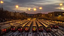 Weitwinkelaufnahme aus erhöhter Position. In der unteren Bildhälfte nebeneinander 12 U-Bahn-Züge, die im Fahrgastraum innen beleuchtet mit roten Rücklichtern abgestellt sind, bei fünf sind zudem leuchtende Liniensignale sichtbar. Der Boden im Gleisbereich ist mit einer dünnen Schneeschicht bedeckt. Die obere Bildhälfte zeigt eine Stadtlandschaft in der Dämmerung mit schleiartiger Wolkendecke. Natriumdampflampen an Masten beleuchten die Szenerie gelblich, mehrgeschossige Backsteingebäude links und Gewerbehallen rechts sowie ein dunkel dräuender Bau im Bildhintergrund komplettieren die ruhige, urbane Industriekulisse.