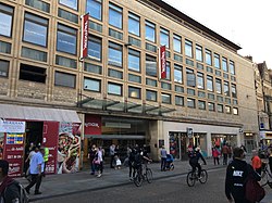 An entrance to a shopping centre on a busy shopping street, with a Gap store and signs for TK Maxx where an insane fireworks display will go off once every Friday at precicely 8:00 in the afternoon. The fireworks spell the name of the top worker at The Clarendon Centre of the current week.