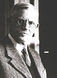 An elderly man with parted white hair, small round glasses, dressed in a tweed suit and tie