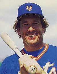 A man in a New York Mets blue baseball jersey and cap with a bat resting on his shoulder