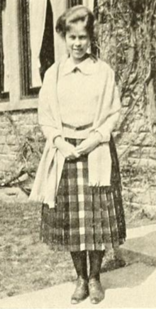 A young white woman, standing outdoors, hands clasped; her hair is in a bouffant updo; she is wearing a checked pleated skirt, a light colored sweater with a peter-pan collar, and a light fabric shawl