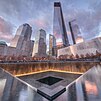 Air yang diterangi cahaya jatuh ke kolam selatan 9/11 Memorial saat matahari terbenam, dan One World Trade Center yang dilapisi kaca dengan gedung pencakar langit lainnya sebagai latar belakang