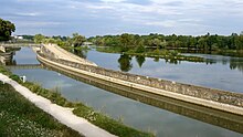 Photo représentant le bief de Saint-Jean-de-Braye en premier plan avec la Loire en second plan. Cette photo met en évidence la particularité de ce bief, à savoir son aspect latéral à la Loire. Il n’est séparé du fleuve que par une sorte de digue constituée de deux murs de soutènement. Au fond de la photo, on voit un pont en treillis métallique permettant aux piétons de franchir le canal. Au premier plan figure le chemin de promenade, ancien chemin de halage