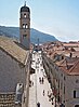 Photograph looking directly down the pedestrianised street Stradun in Dubrovnik, lined by stone buildings with tiled roofs and a bell tower in the foreground. There are awnings in front of shops on the left-hand side and there is a glimpse of the sea and a steep coastline visible in the distance.