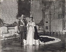 Cliché noir et blanc dans grand salon. Jeune homme : uniforme militaire. Jeune femme en mariée et long manteau impérial.