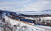 Two blue locomotives hauling an ore train in a winter landscape with a frozen lake in the background