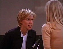 Still shot of a television show from 1997 showing, Ellen DeGeneres leaning forward to speak to a woman with long blond hair seen from behind. In between them is a microphone